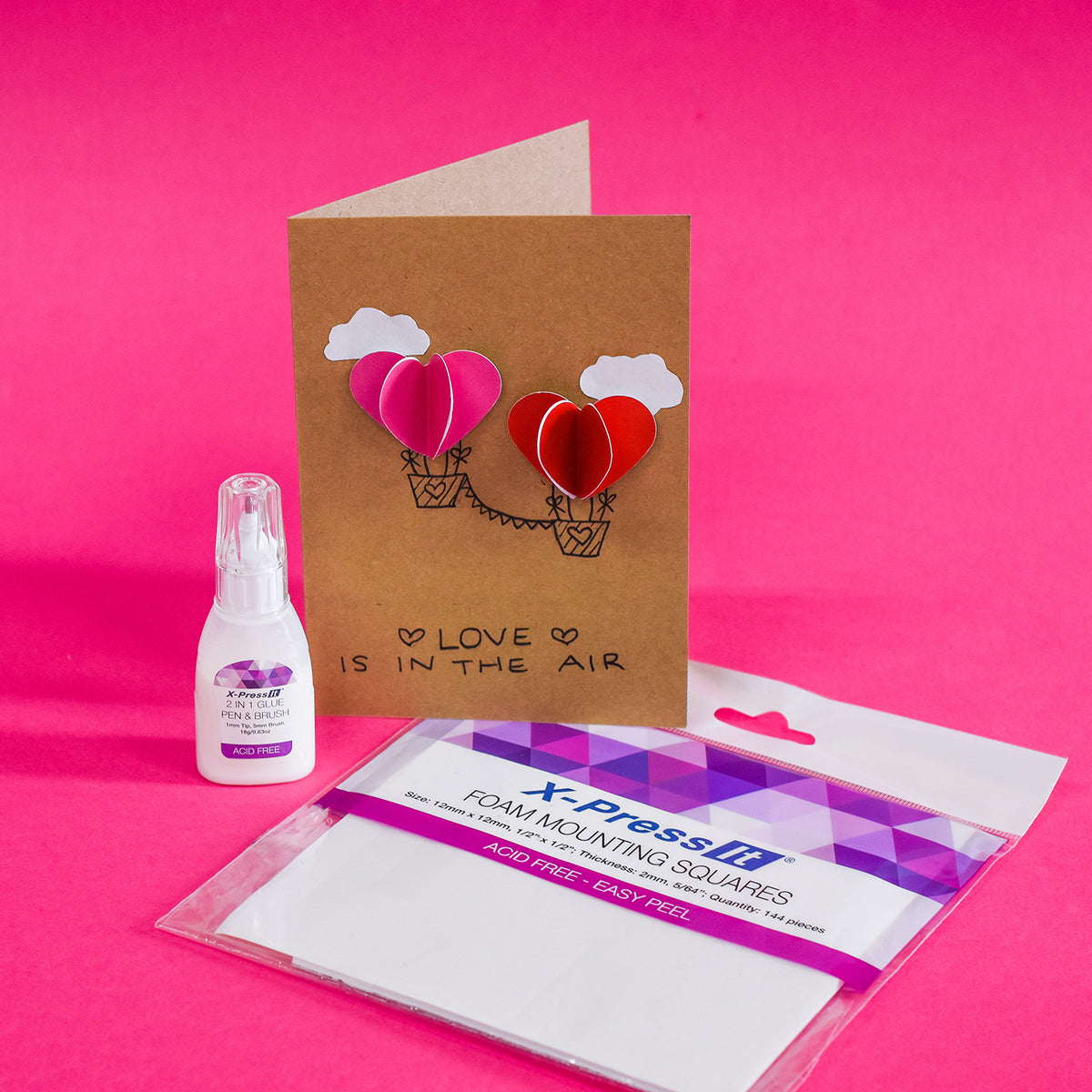 Kraft card with 3D pink and red paper heart balloons, a glue pen bottle and the foam squares pack on a pink background