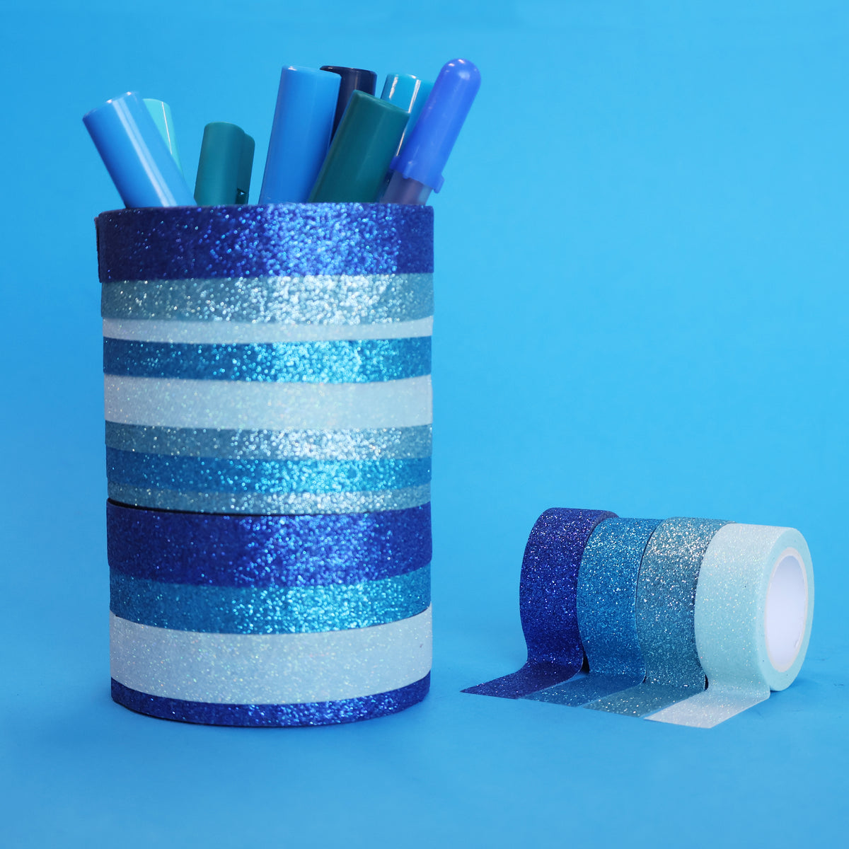 Pen pot striped in blue glitter tape holding blue pens, with the four X-Press It blues rolls beside it
