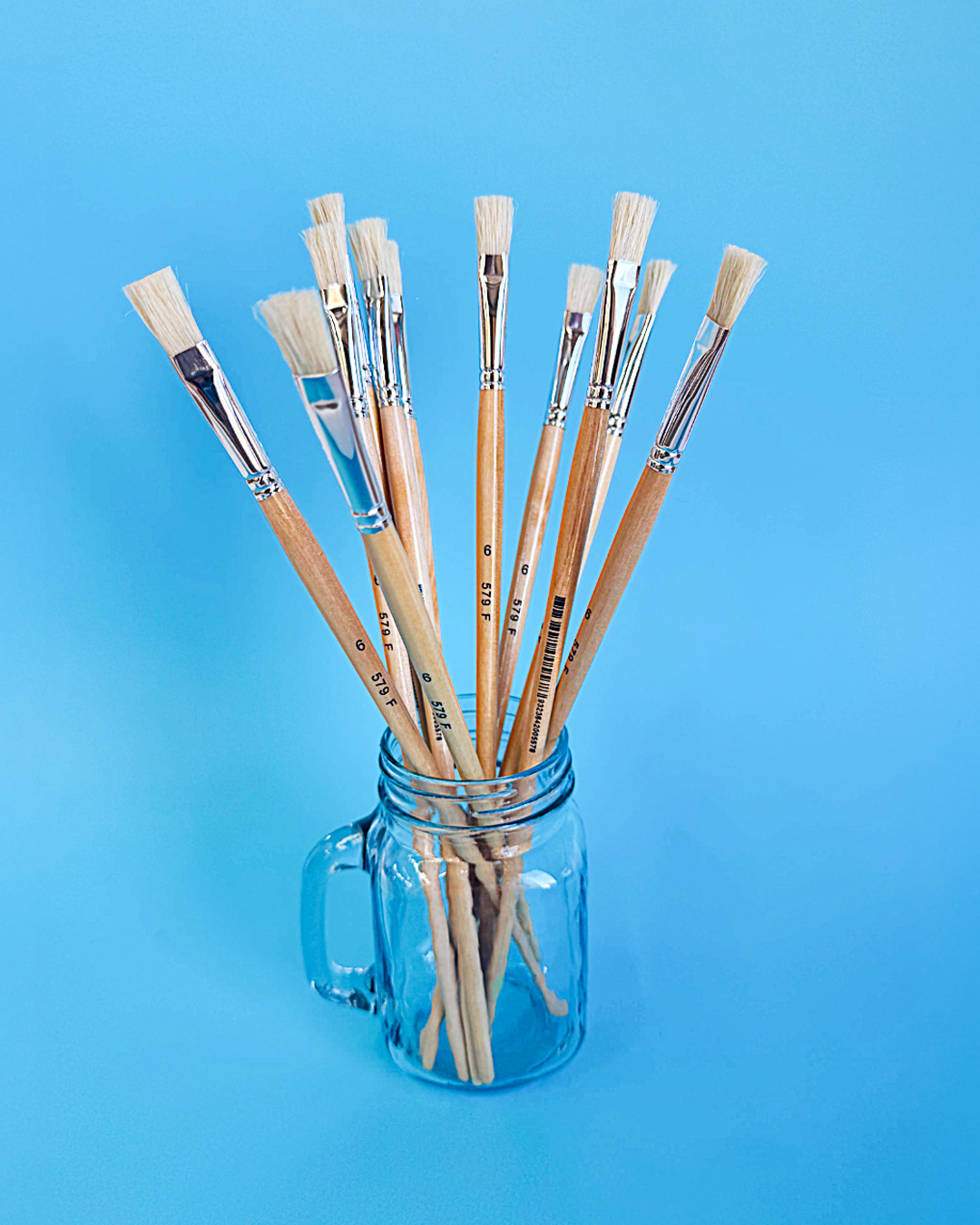 Assorted X-Press It hog hair flat and round brushes standing in a glass jar on a bright blue background, in a glass mug jar