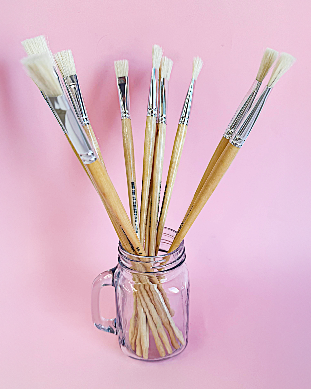 Assorted X-Press It hog hair flat and round brushes standing in a glass jar on a soft pink background, in a glass mug jar