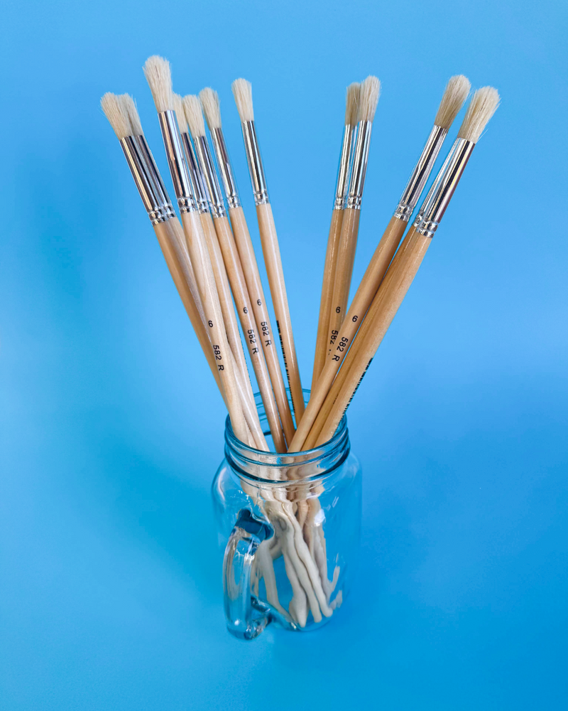 Glass mug filled with X-Press It size 6 hog hair round brushes, bristles up, on a bright blue background