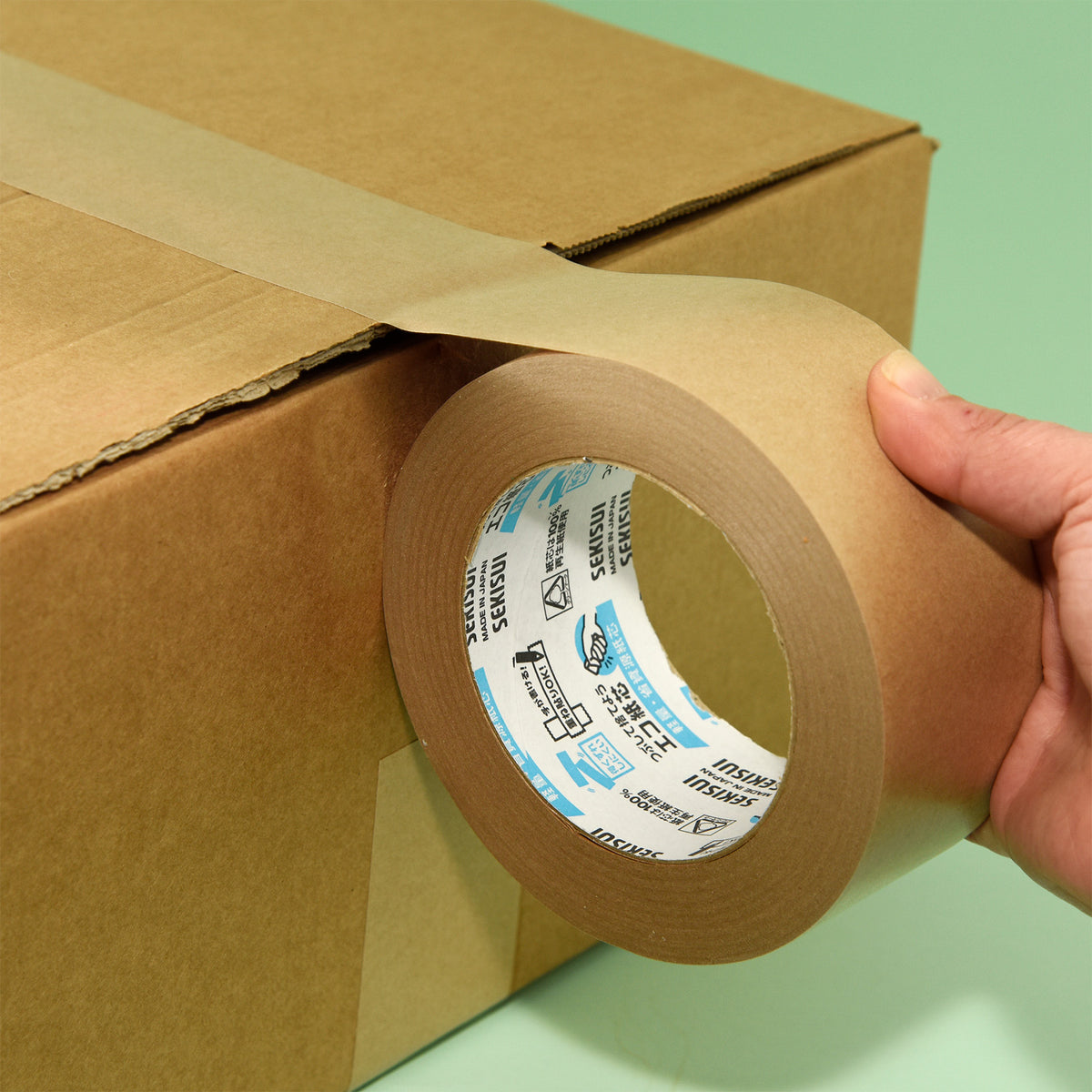 Hand laying wide brown kraft tape across a cardboard box seam against a green background