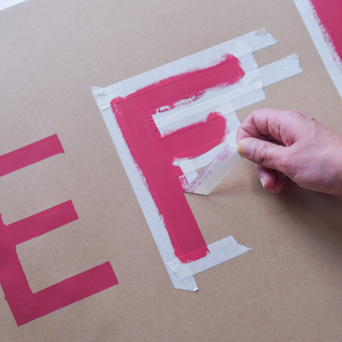 Hand peeling masking tape away from painted pink block letters on brown board
