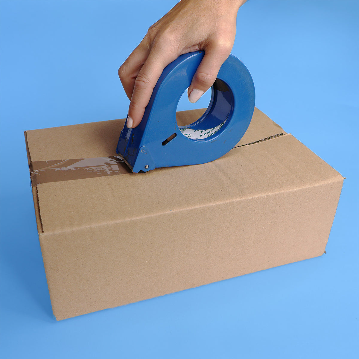 Hand using a blue tape dispenser to seal the seam of a cardboard box with brown tape on a blue background