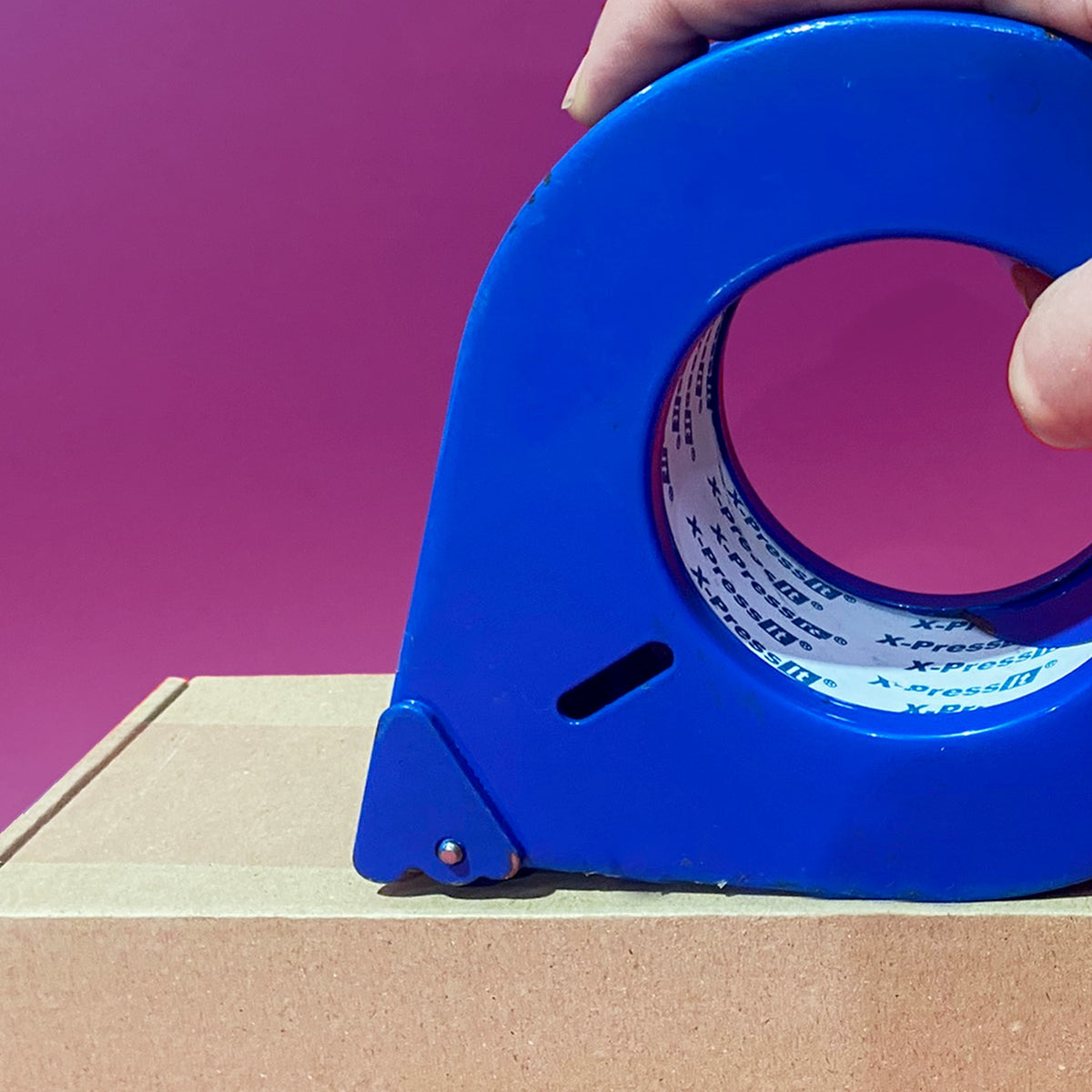 Hand pressing a blue tape dispenser along the edge of a cardboard box against a magenta background
