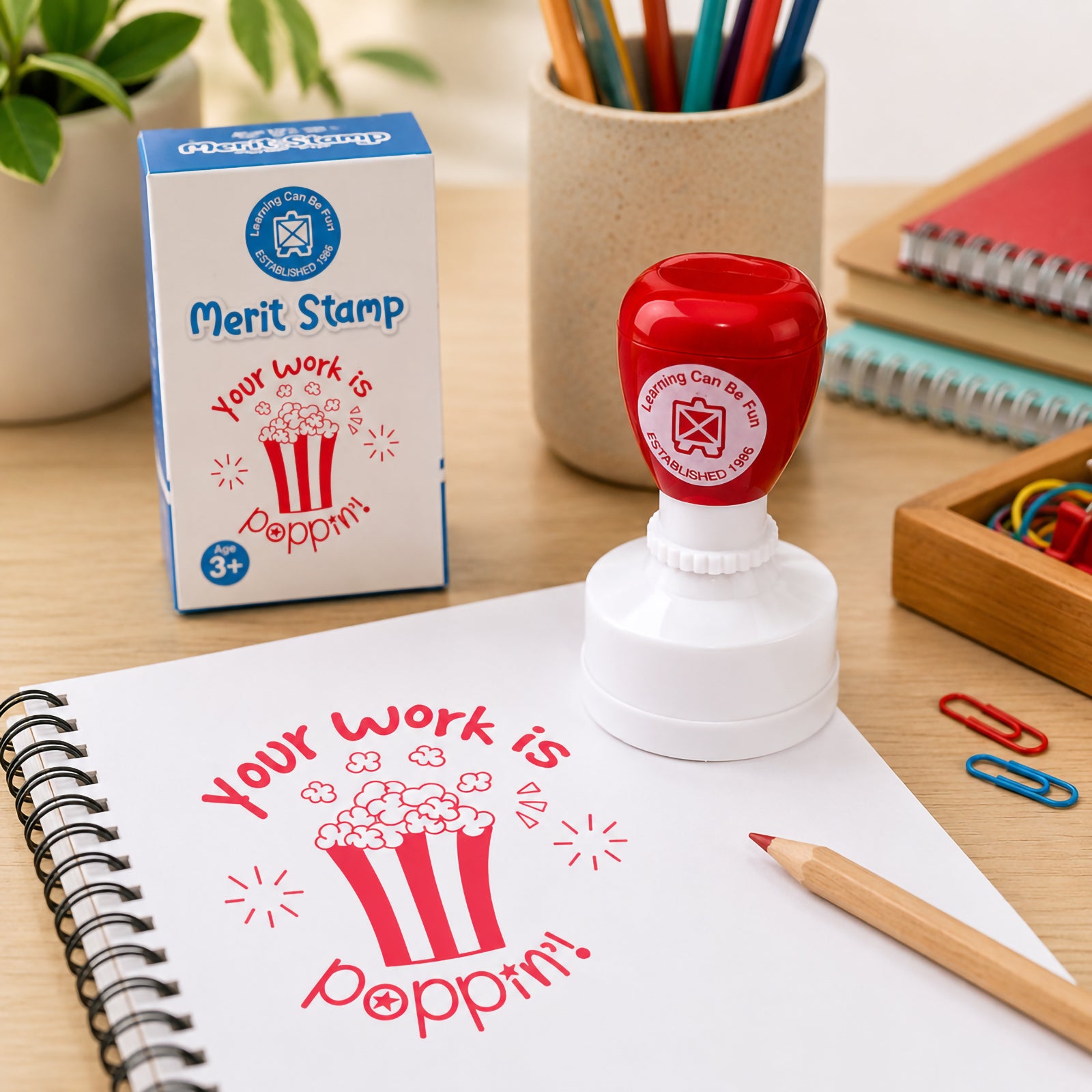 Your Work Is Poppin Merit Stamp positioned next to its retail packaging on a classroom workspace. The image showcases the bold popcorn design, red ink impression and practical teacher reward stamp used for recognising achievement and encouraging learners.