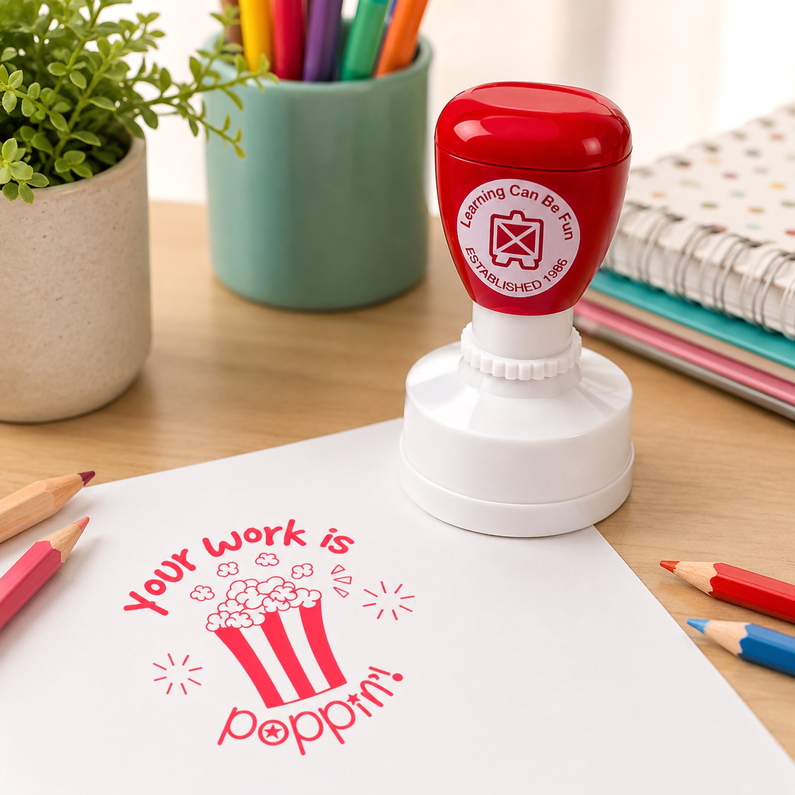 Self inking merit stamp standing on a classroom desk beside coloured pencils, notebooks and stationery. A vibrant red Your Work Is Poppin impression is stamped onto paper, demonstrating how teachers can provide positive feedback and recognise student effort.