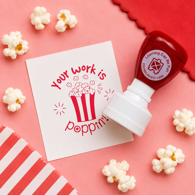 Learning Can Be Fun Your Work Is Poppin Merit Stamp displayed on a pink background surrounded by popcorn pieces. The red self inking teacher stamp sits beside a bright popcorn themed impression, highlighting a fun and engaging classroom reward tool designed to celebrate student achievement.