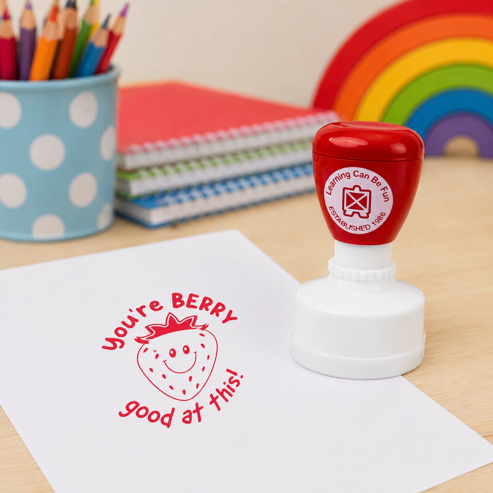 Red merit stamp positioned beside a worksheet displaying the cheerful strawberry design and encouraging message. Classroom supplies and a colourful rainbow backdrop create an inviting educational setting that showcases rewarding achievement and effort.