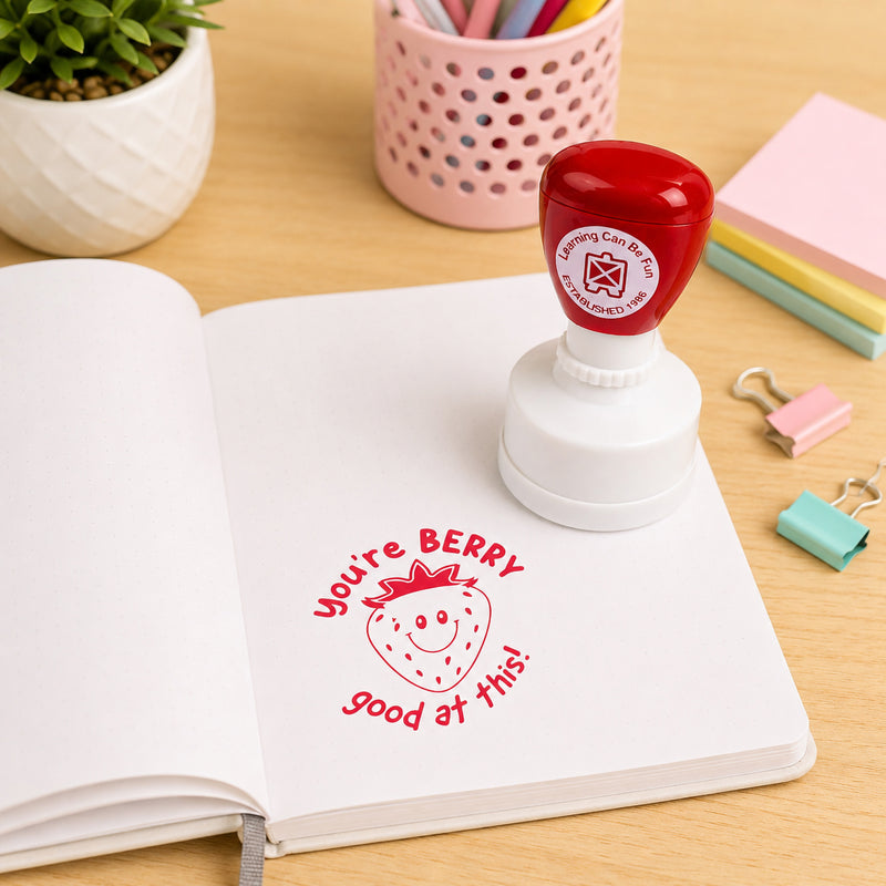 Learning Can Be Fun You're Berry Good At This Merit Stamp shown beside an open notebook featuring a bright red strawberry themed stamp impression. The self inking reward stamp is displayed in a classroom inspired workspace with stationery and colourful accessories, highlighting its use for positive student recognition.