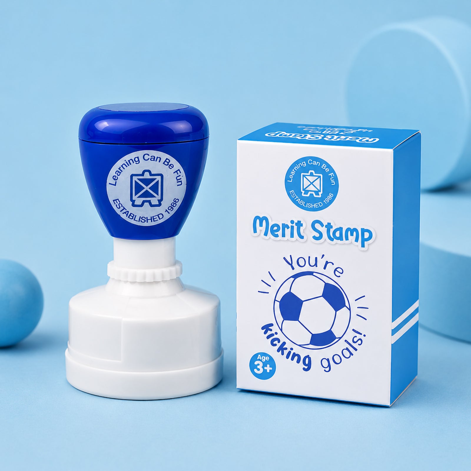 Product packaging displayed alongside the merit stamp on a blue background. The packaging showcases the soccer ball artwork and motivational message, presenting the stamp as an inspiring classroom reward tool.