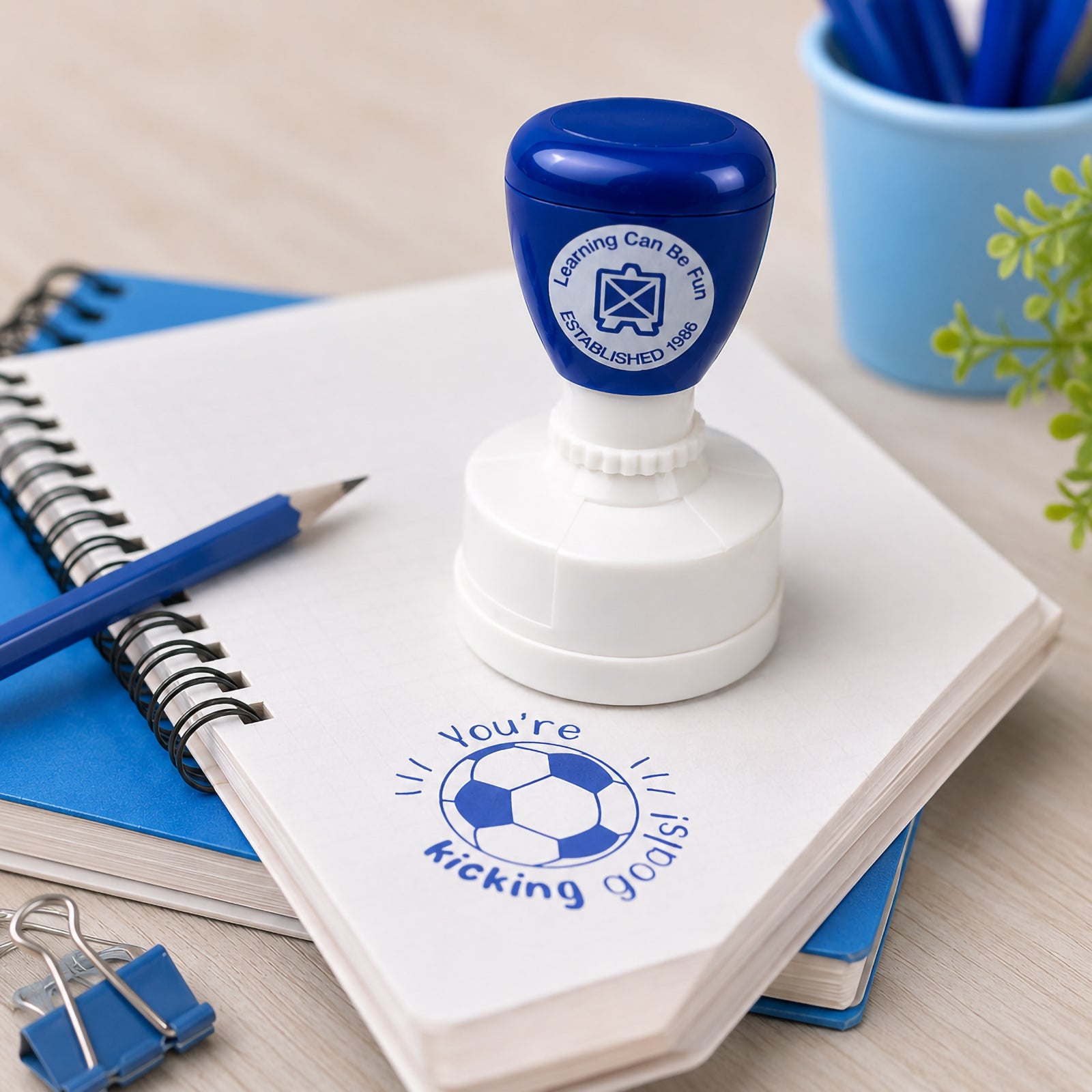 The self inking merit stamp stands on an open notebook beside blue stationery and classroom supplies. A clear You're Kicking Goals soccer themed impression demonstrates the bold 36mm design and vibrant blue ink, ideal for recognising effort and achievement.