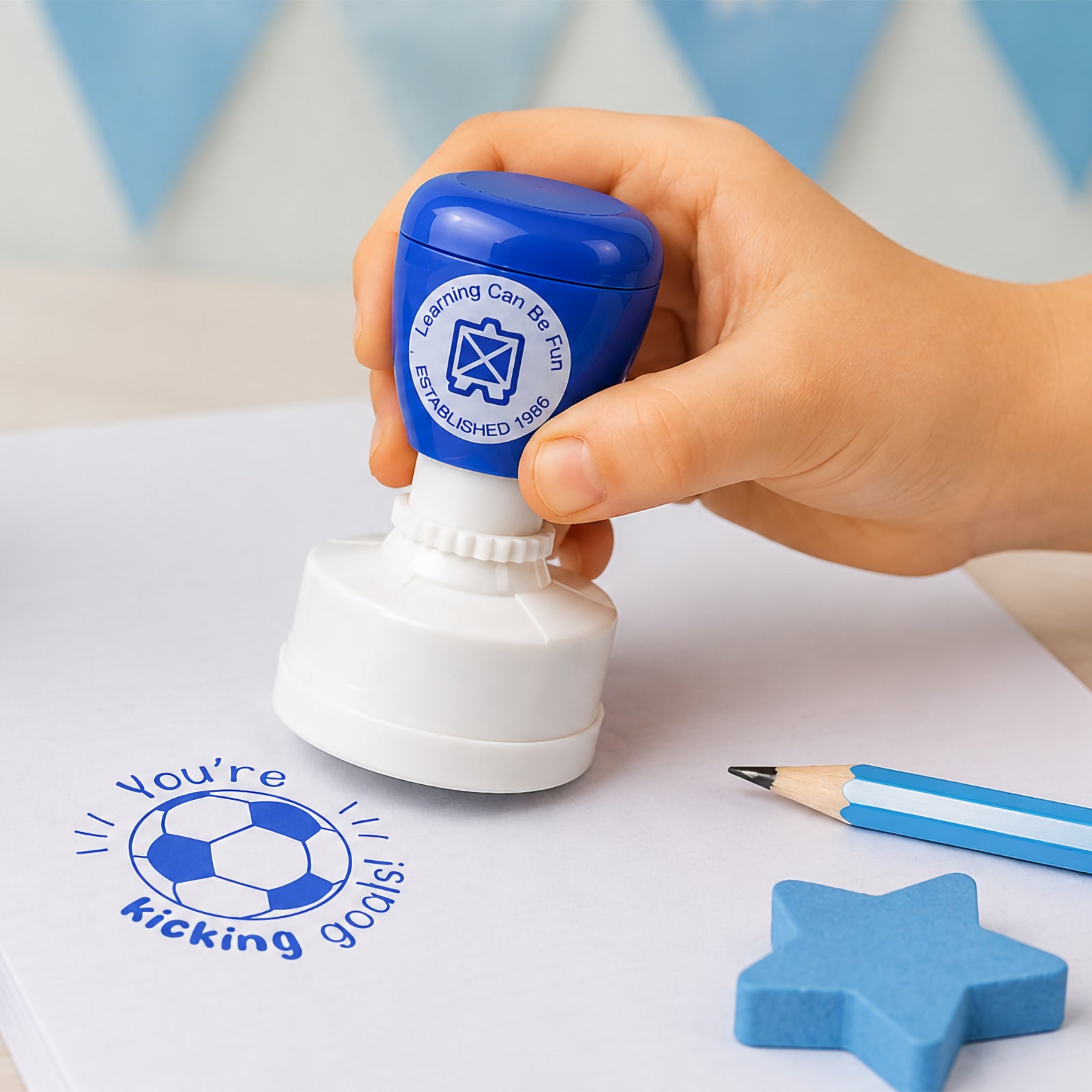 A child uses the Learning Can Be Fun You're Kicking Goals Merit Stamp on a sheet of paper, creating a crisp blue soccer ball impression with the encouraging message. The image highlights the easy self inking operation and classroom friendly design that makes rewarding student achievement simple and engaging.