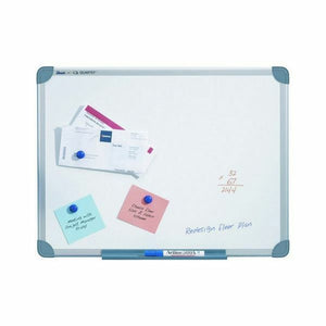 Quartet commercial whiteboard in an aluminium frame with grey corner caps and a small printed label on the top rail, printed papers under a blue magnet, a blue note, a pink note and a marker in the tray