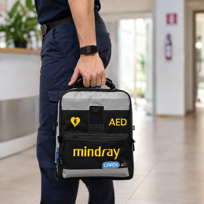 CardiAct Mindray Soft Carry Case being carried by its reinforced top handle through a bright workplace or public facility. The black and grey AED bag features prominent yellow AED and Mindray identification with CardiAct branding, demonstrating convenient grab and go transport for rapid emergency response.