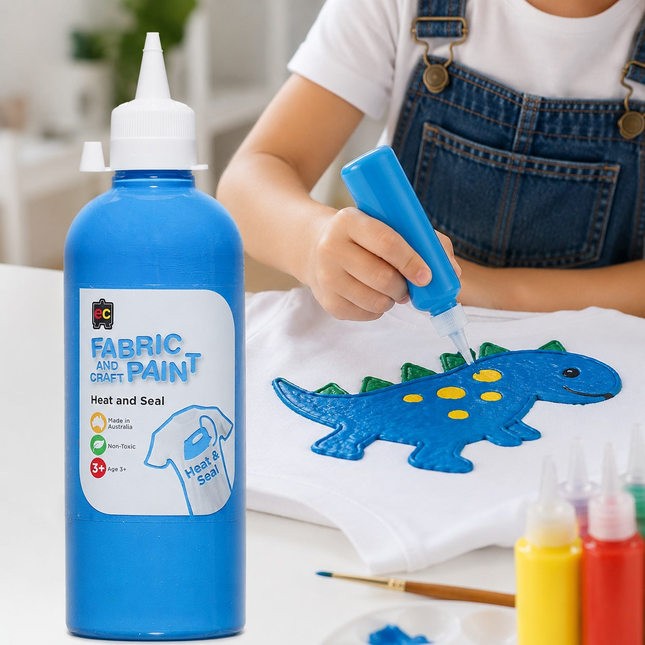 Child painting a dinosaur design on fabric with blue fabric paint, next to a bottle of fabric paint.