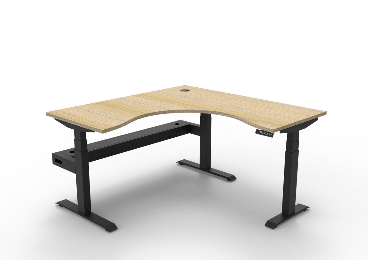 Rapidline Boost+ corner sit stand workstation, Natural Oak top on black triple motor frame and cable tray underneath, front three-quarter view