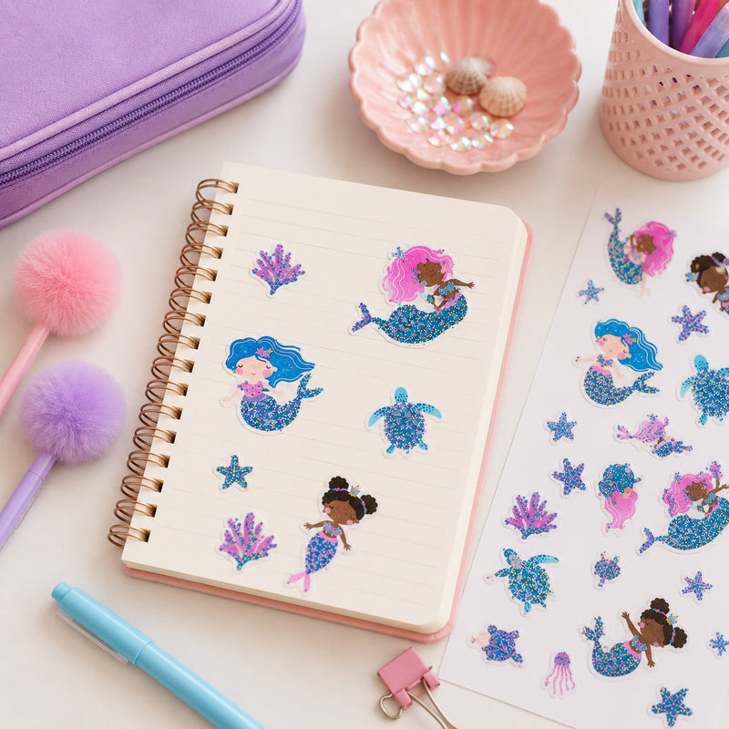 Mermaid Magic sticker sheet displayed beside a notebook, fluffy pens and decorative seashell accessories. The glitter accented collection features mermaids, turtles, coral and ocean themed characters arranged for creative crafting inspiration.