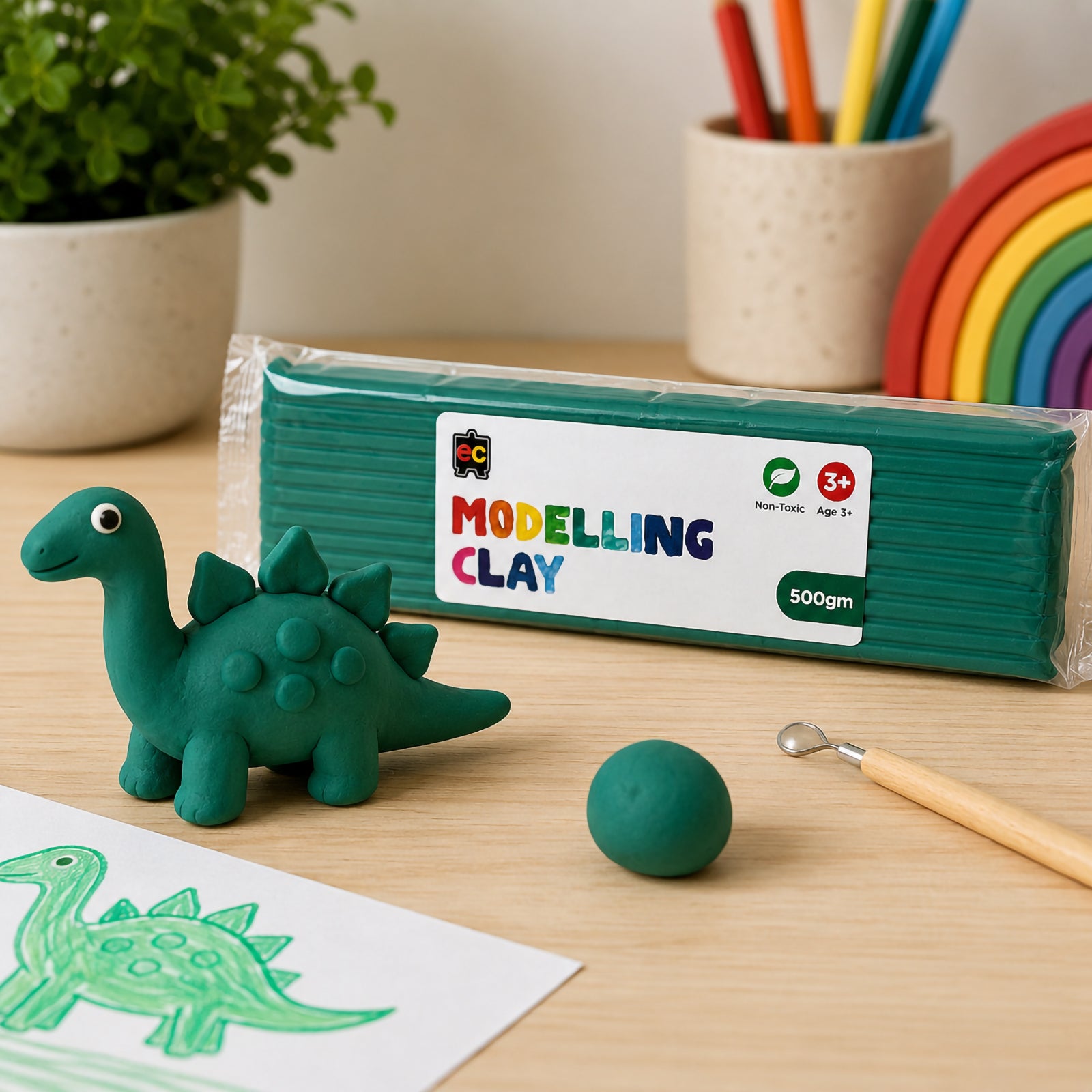Educational Colours 500g dark green modelling clay pack beside a handmade clay dinosaur sculpture, rainbow toy, drawing sketch, and craft tools on a clean wooden desk with colourful art supplies in the background.