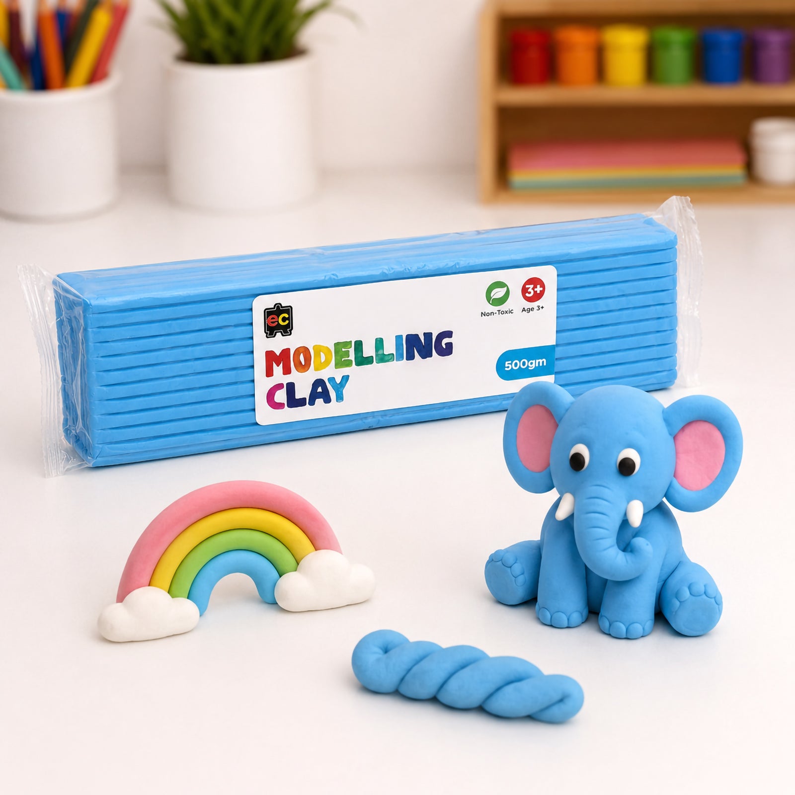 Educational Colours light blue modelling clay pack beside a clay elephant, rainbow and twisted clay rope on a clean white craft table with colourful supplies in the background.