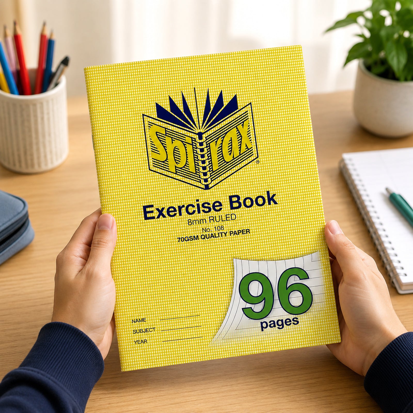Hands holding a Spirax P138 A4 Graph Book with a bright yellow cover on a tidy study desk surrounded by stationery, notebook and indoor plants, highlighting the durable design and large 10mm graph format for everyday school use.
