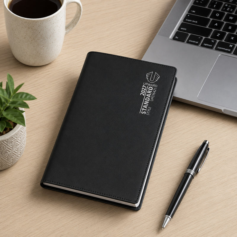 The closed Upward Standard 2027 diary rests on a light timber desk next to a laptop, coffee mug and pen. Its black PVC cover presents a clean professional appearance while the compact A6/7 format makes it ideal for portable planning and organisation.