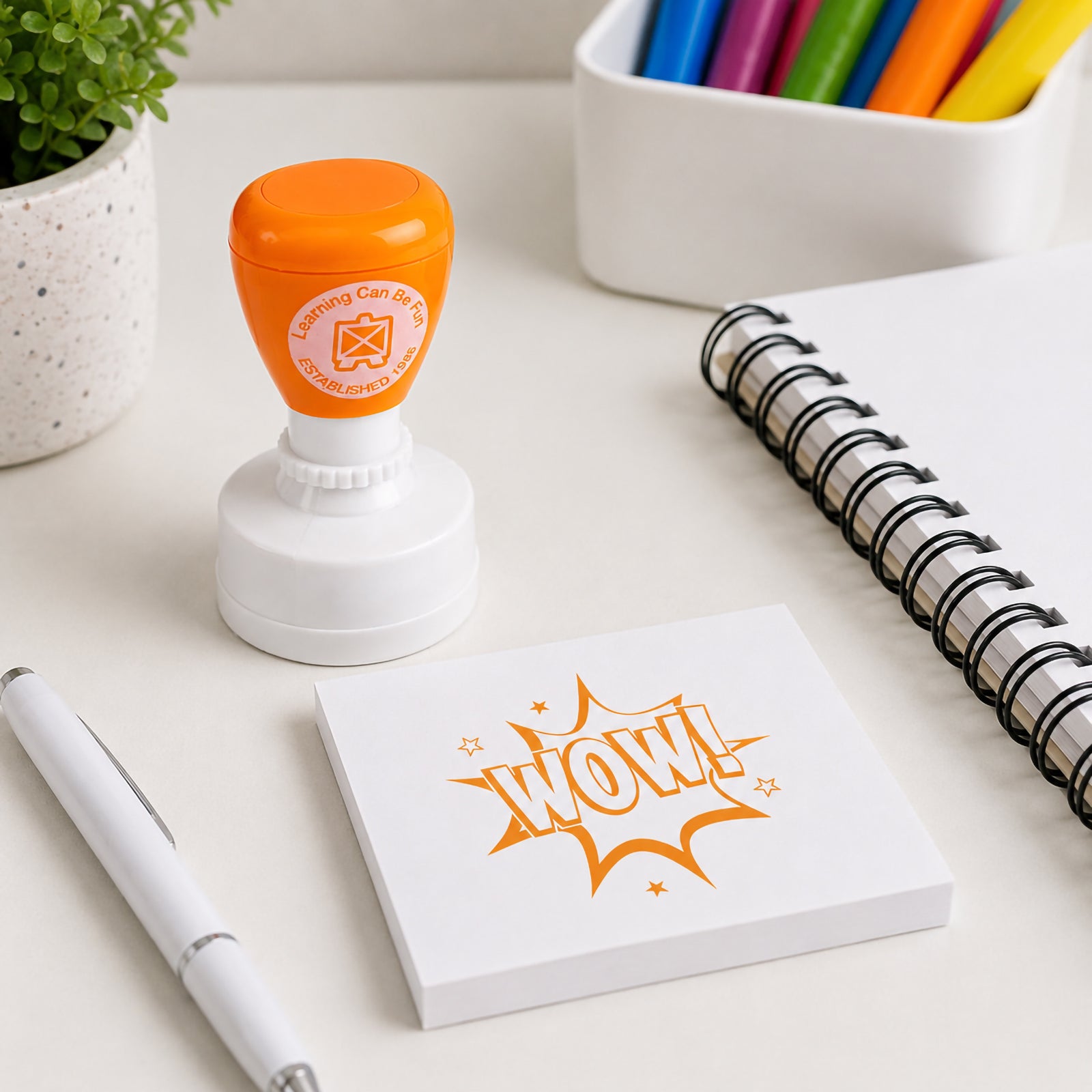 Learning Can Be Fun Wow Merit Stamp displayed on a desk beside a memo pad featuring a bold comic style WOW! stamp impression. The bright orange self inking stamp is shown with classroom stationery, highlighting its use for rewarding student achievement and recognising excellent work.
